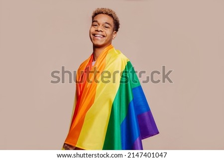 Portrait of happy smiling afro latin american young man with a gay pride rainbow flag at studio over beige background. Homosexual lgbtiq concept, rainbow flag, celebrating parade. Copy space. Royalty-Free Stock Photo #2147401047