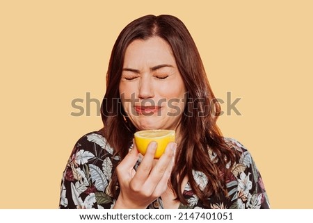 Face portrait of woman wears floral shirt, eating acid lemon doing funny face expression with eyes closed. Studio shot over yellow background.  Royalty-Free Stock Photo #2147401051