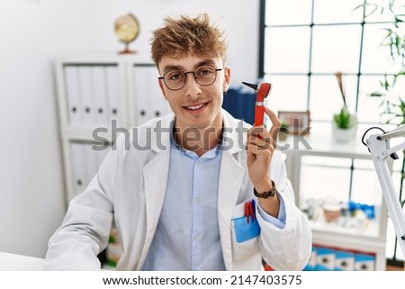 Young caucasian doctor man holding ear otoscope at the clinic looking positive and happy standing and smiling with a confident smile showing teeth 