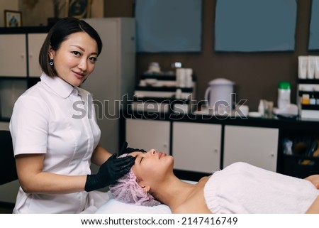 Female beautician doing massage of girl face and looking at camera in modern beauty salon. Young asian pretty woman client with closed eyes wearing medical cap. Concept of body and skin care