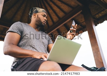 African American male using cellphone device for sharing media files to laptop computer connecting to 4g wireless and bluetooth, millennial graphic designer with netbook chatting via mobile app