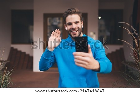 Millennial hipster guy in earbuds enjoying video calling during leisure pastime in city, cheerful male blogger in electronic headphones creating selfie content clicking photos on web camera