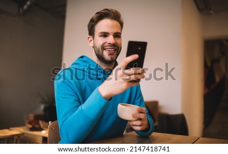 Cheerful hipster guy with coffee cup enjoying mobile messaging in social media using cafeteria wifi connection on weekend, happy Caucaisan blogger checking text sms while making online booking