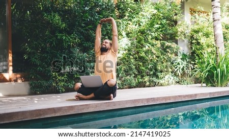 Horizontal view of Caucasian IT professional stretching hands while doing distance job - creating graphic design for web startup project, millennial male programmer with netbook at pool terrace