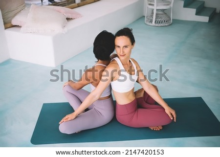 Girlfriends practicing yoga in Twist pose at home. Concept of healthy lifestyle. Idea of friendship. Women wearing sportswear sitting back to back on fitness mat. caucasian girl looking at camera