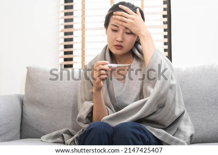 Sick, influenza asian young woman, girl headache have a fever, flu and check thermometer measure body temperature, feel illness sitting on sofa bed at home. Health care person on virus, covid-19. Royalty-Free Stock Photo #2147425407