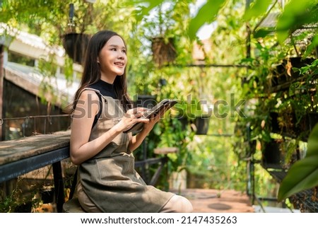 Asian female Woman gardener in apron working in a outdoor garden at home studio.Female Gardener using tablet computer for setting water drop system in greenhouse,modern greenhouse farm ideas concept