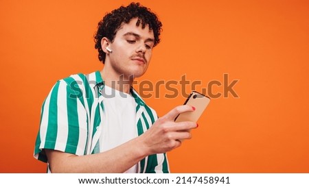 Surfing the net for vibrant music. Confident queer man using a smartphone while wearing wireless earphones. Non-conforming young man listening to his favourite playlists against an orange background.