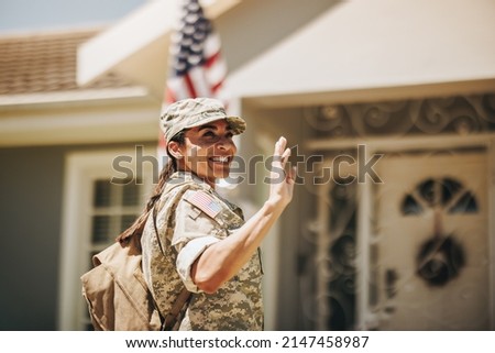 Happy female soldier smiling and waving her hand proudly while standing outside her home. Patriotic American servicewoman coming back home after serving her country in the military. Royalty-Free Stock Photo #2147458987