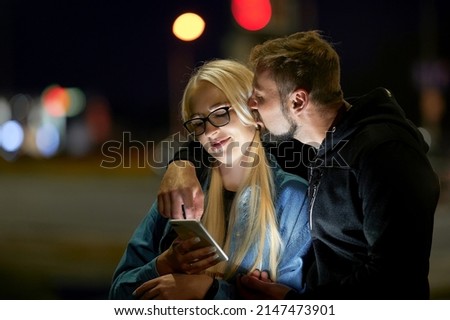 A beautiful couple stands and hugs at night and looks into the phone. Date, he kisses on the ear.