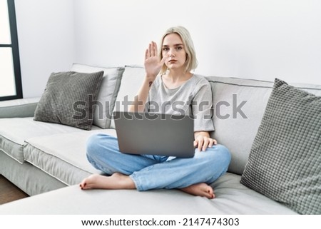 Young caucasian woman using laptop at home sitting on the sofa doing stop sing with palm of the hand. warning expression with negative and serious gesture on the face. 