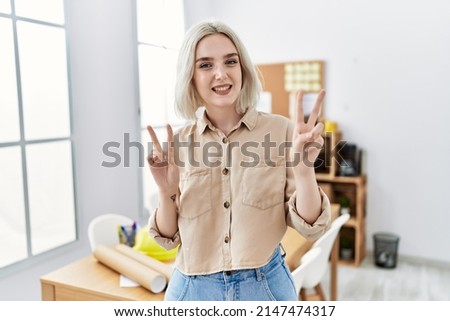 Young beautiful caucasian woman at construction office smiling looking to the camera showing fingers doing victory sign. number two. 