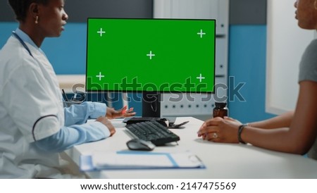 Close up of computer with horizontal green screen on desk. Doctor and patient sitting and looking at monitor with chroma key and isolated background or mockup template on display.