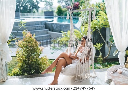 Young caucasian woman sitting and resting on hammock chair on terrace of resort hotel. Concept of tourism, vacation and weekend. Smiling girl with tattoos holding glass cup. Sunny day. Bali island Royalty-Free Stock Photo #2147497649