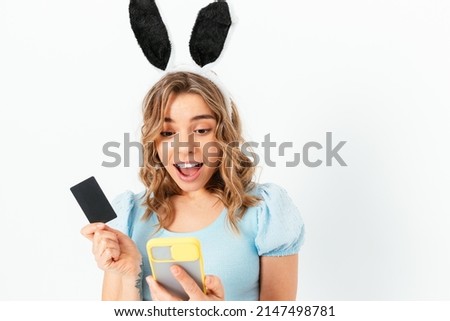 Delighted young woman wearing Easter Bunny ears holding smartphone and credit card in her hands, white studio background