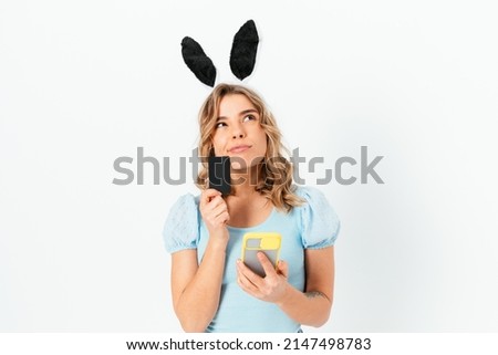 Pensive young woman wearing Easter Bunny ears makes online purchases through her smartphone holding credit bank card in her hand looks up thoughtfully