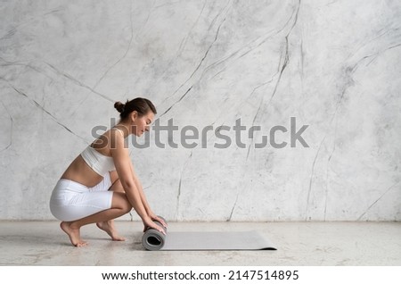 Woman rolling her gray mat on the floor with both hands on after yoga class with copy space on the wall