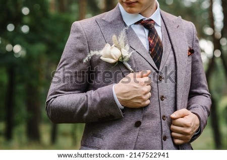 The groom with tie in a suit with boutonniere or buttonhole on jacket, is stands on the background greenery in the garden, park. Nature. Cropped photo.