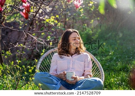 Young beautiful woman enjoying relaxing in the spring while sitting in the blooming garden of her home with a cup of tea Royalty-Free Stock Photo #2147528709