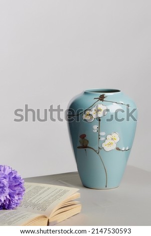 Shot of a blue hand-painted glossy vase made of ceramic with a sakura pattern standing on a grey table. The flower and book are lying next to the vase. The glazed vase is isolated against grey wall.