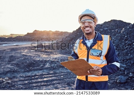 A young African mine worker wearing protective wear is looking at the camera while holding a clipboard with coal mine in the background Royalty-Free Stock Photo #2147533089