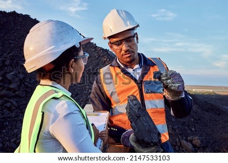 Two young African mine workers wearing protective wear are discussing coal while on site at a coal mine Royalty-Free Stock Photo #2147533103