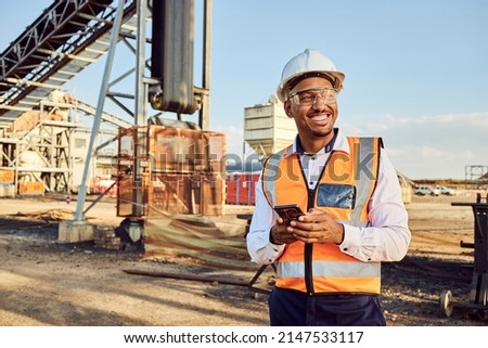 A young African mine worker wearing protective wear is looking off camera while holding a cell phone with coal mine equipment in the background Royalty-Free Stock Photo #2147533117