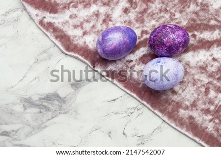 Three purple eggs lie on a velvet cloth on a marble background. Easter card with a place for text in pantone colors