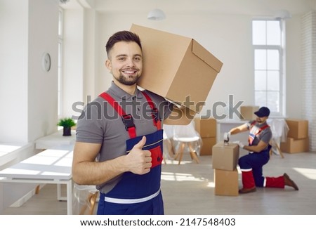 Smiling male worker of moving and delivery company holding cardboard box showing thumbs up. Loader in overalls posing against background of colleague who packs cardboard boxes. Moving service concept.