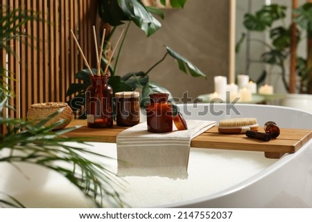 Wooden bath tray with open book, candle and body care products on tub indoors. Relaxing atmosphere