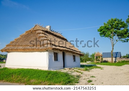 Rural landscape with ancient Ukrainian clay house. Thatched roof, white walls with lime, chimney. National Ukrainian traditions. Royalty-Free Stock Photo #2147563251