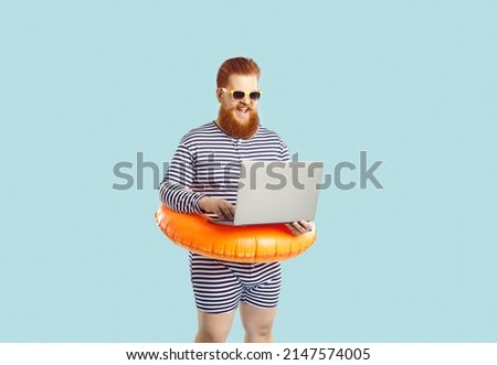 Work and vacation. Funny excited chubby man on pastel turquoise background working remotely with laptop during summer vacation. Red-bearded man in striped leotard with inflatable circle at waist. Royalty-Free Stock Photo #2147574005