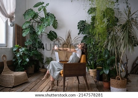 Overworked woman freelancer sitting on chair in cozy greenhouse, resting, stretching arms with closed eyes. Smiling tired woman in home garden taking break from online study. Plant lovers concept. Royalty-Free Stock Photo #2147578281