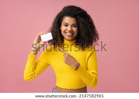 Beautiful smiling curly haired woman holding blank business card pointing finger on copy space isolated on pink background. Mockup, advertisement concept 