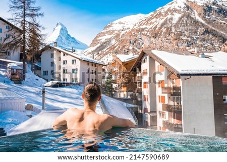 Tourist swimming in pool while looking at matterhorn mountains and houses. Rear view of man relaxing in hot tub. View of snow covered landscape in alps during winter.