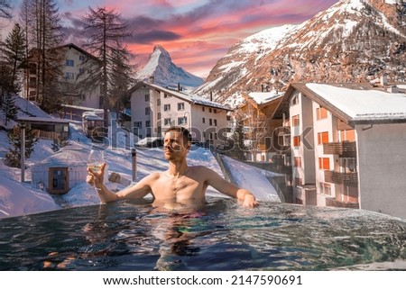 Man enjoying wine while swimming in pool against townscape. Tourist relaxing in hot tub against matterhorn mountain. Scenic view of snow covered landscape during sunset.