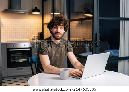 Portrait of cheerful bearded young man using typing on laptop sitting at table in kitchen room with modern interior, looking at camera. Confedent curly happy freelancer male remote working from home. Royalty-Free Stock Photo #2147603385