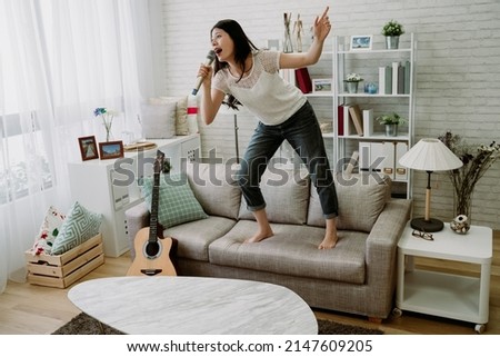 weekend day asian girl is standing on couch blowing off steam by singing karaoke at home. chinese woman holding microphone with exaggerated body movement is having happy and crazy time alone. Royalty-Free Stock Photo #2147609205