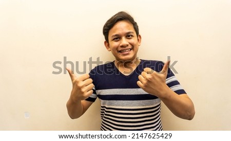 Wow and impressed face reaction of young Asian Malay man with two thumbs up and strips t-shirt on isolated white background. Selective focus.