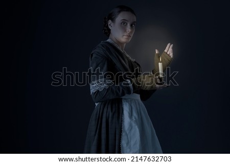 A working class Victorian woman wearing a dark green checked bodice and skirt and holding a candle in a darkened room Royalty-Free Stock Photo #2147632703