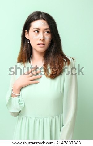 Young Asian woman with glitter makeup touching necklace on color background