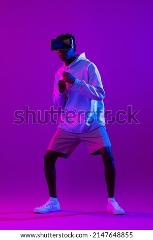 Young African man wearing VR glasses and interacting with virtual reality game in modern neon light studio background