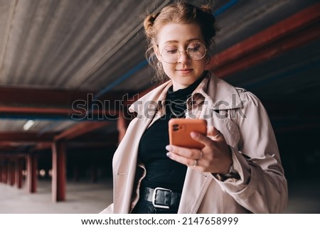 young caucasian female student, generation z, dressed in a trench coat in the parking lot uses a smartphone Royalty-Free Stock Photo #2147658999