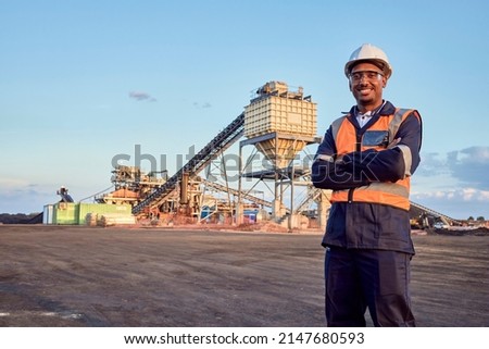 A young African mine worker wearing protective wear is looking at the camera with coal mine equipment in the background Royalty-Free Stock Photo #2147680593