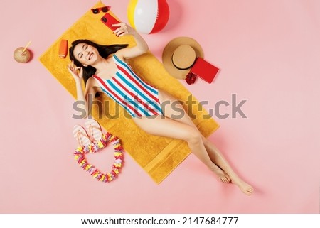 Top view full body young woman of Asian ethnicity in swimsuit lies on towel hotel pool do selfie shot mobile cell phone show ok isolated on plain pastel pink background. Summer vacation sea concept