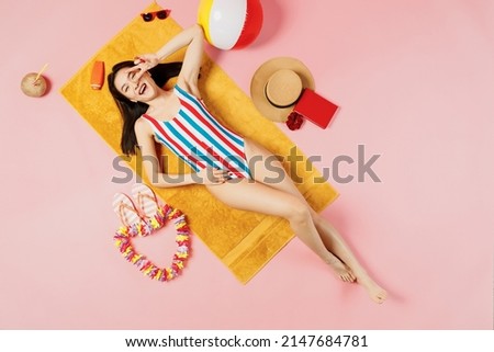 Top view full body young fun happy woman of Asian ethnicity in striped swimsuit lies on towel hotel pool show v-sign isolated on plain pastel pink background. Summer vacation sea rest sun tan concept