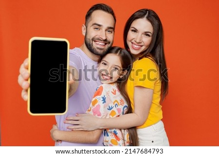 Young happy parents mom dad with child kid daughter teen girl in basic t-shirts hold use mobile cell phone with blank screen workspace area isolated on yellow background studio. Family day concept.