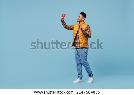 Full size body length young black man 20s wears yellow waistcoat shirt do selfie shot on mobile cell phone post photo on social network isolated on plain pastel light blue background studio portrait