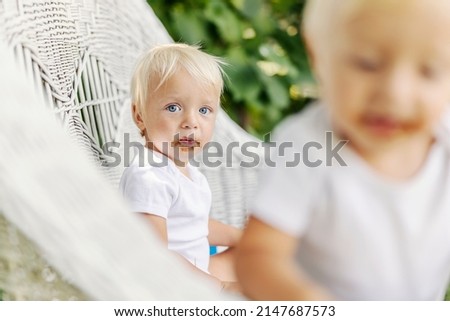 A toddler with blue eyes and blond hair with a dirty chocolate face sits on wicker chairs in the yard and looks at the camera. A cute baby enjoys sweets. Funny picture of a baby