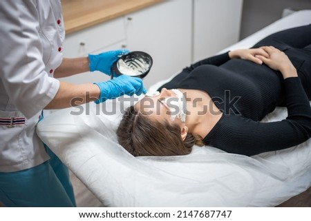 Doctor cosmetologist or dermatologist making face mask in cosmetology salon. Professional Beautician applying face mask on caucasian woman face lying on bed in bathrobe. High quality photo